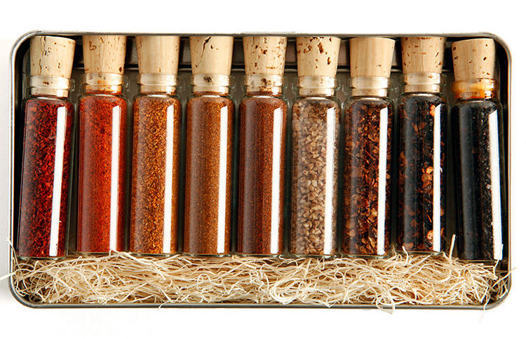 9 glass vials of hot pepper powders and spices, sealed with a cork. In a metal tin with straw packing.